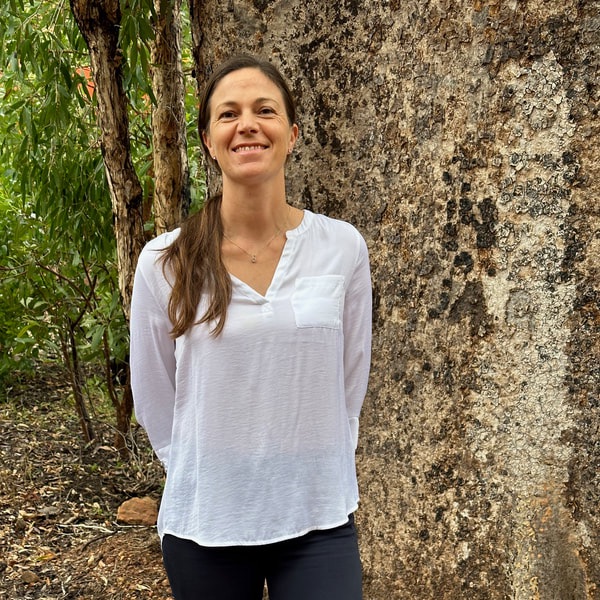 Rebecca is standing in front of a Boab tree, she's wearing a white cotton shirt and navy blue trousers, she's smiling at the camera